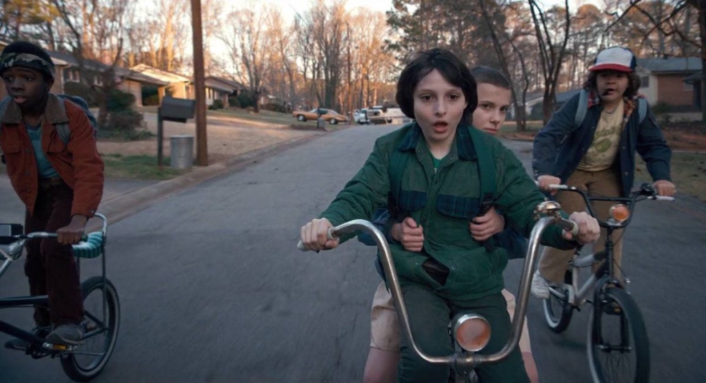 (from left to right) Lucas, Mike, Eleven, and Dustin from Stranger Things riding their bikes down a street.