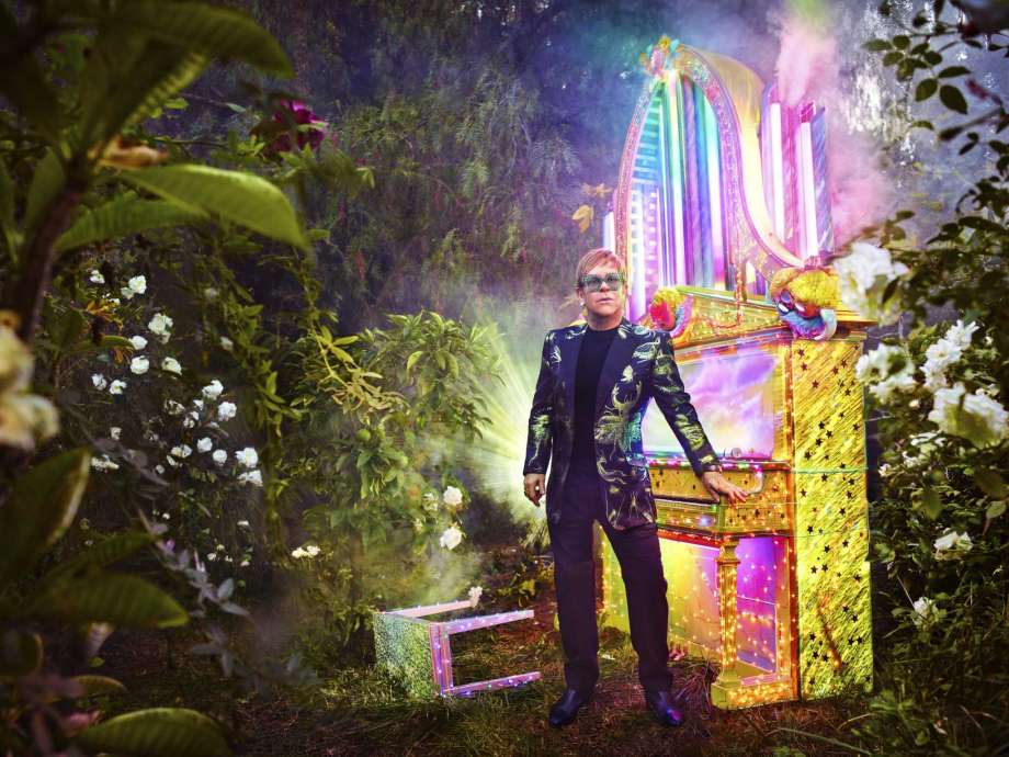 Elton John standing beside a colorfully-lit piano in a forest