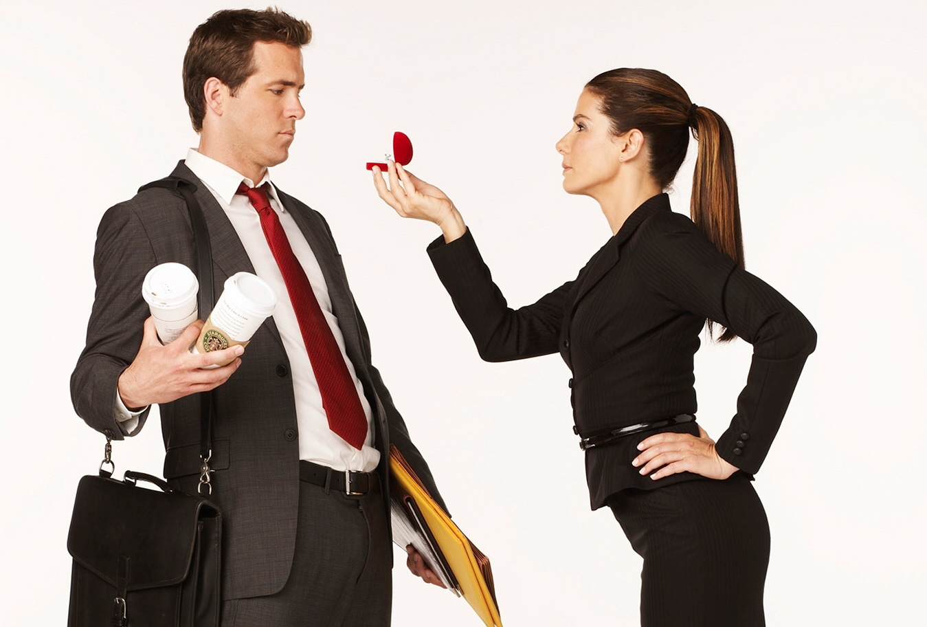 The female lead holds an engagement ring box in front of the male lead's face as he leans away.