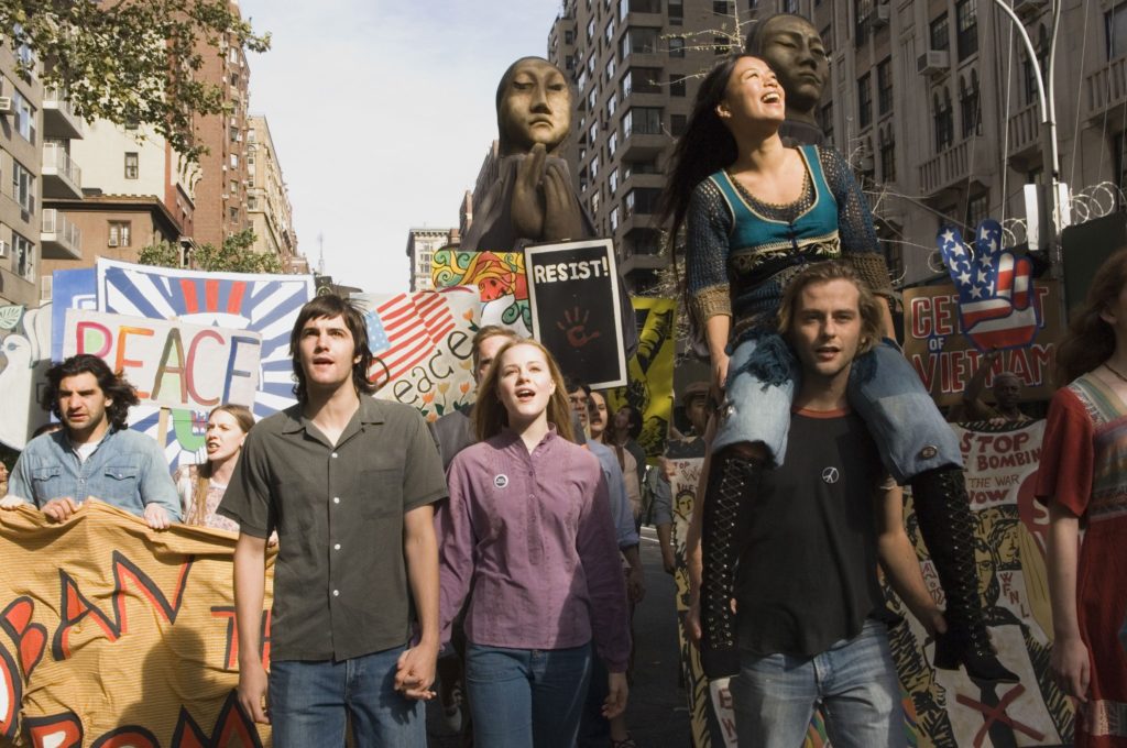 Several characters participate in a march in protest of the Vietnam War. Many signs calling for peace and unusual puppets are seen in the background of this still from the musical. 