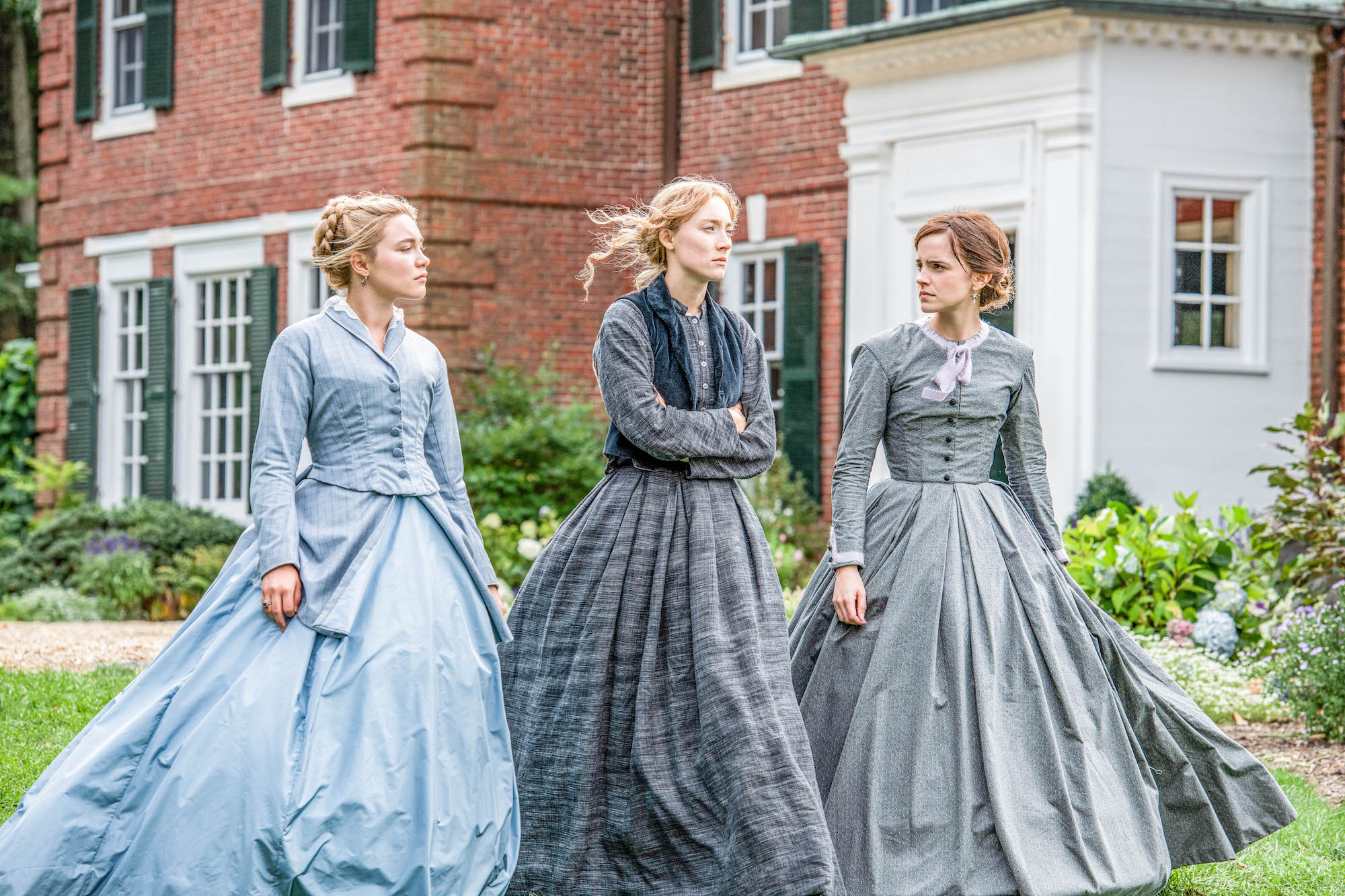 Meg, Jo, and Amy stand in a yard, wearing grey and blue dresses.