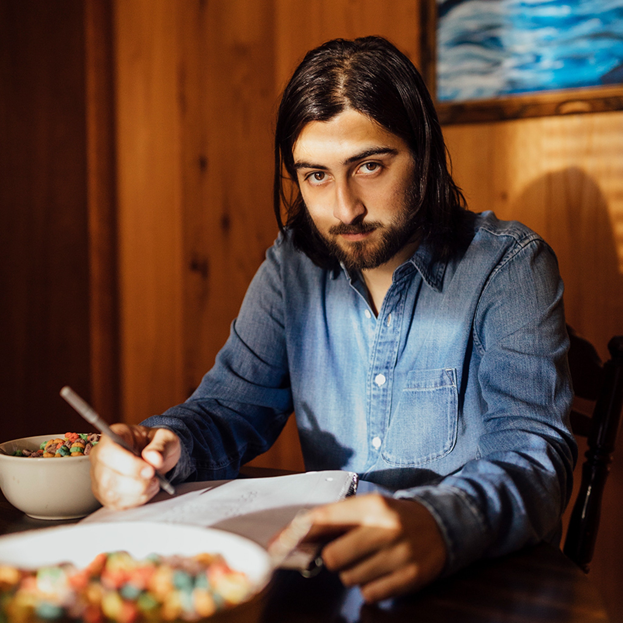 Noah Kahan sitting at a table writing in a notebook while looking at the camera.