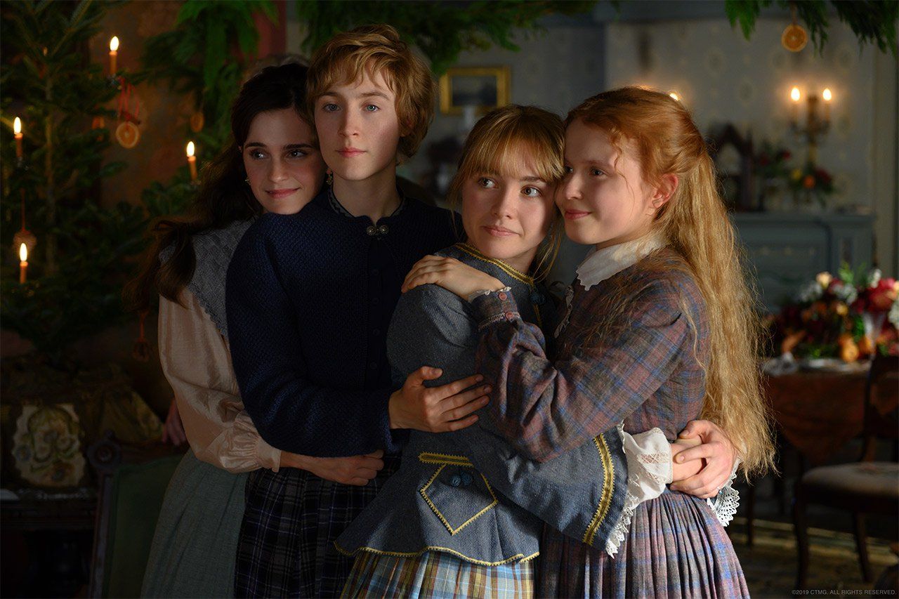 The little women (Amy, Jo, Beth, and Meg) stand in their living room at Christmas, holding each other.