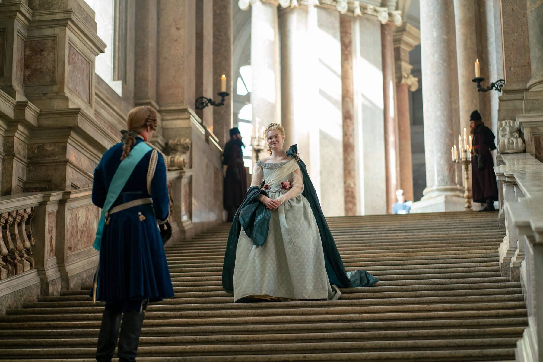 Elle Fanning as Catherine the Great stands on the stairs of the royal palace facing her husband Peter the Third portrayed by Nicholas Hoult.