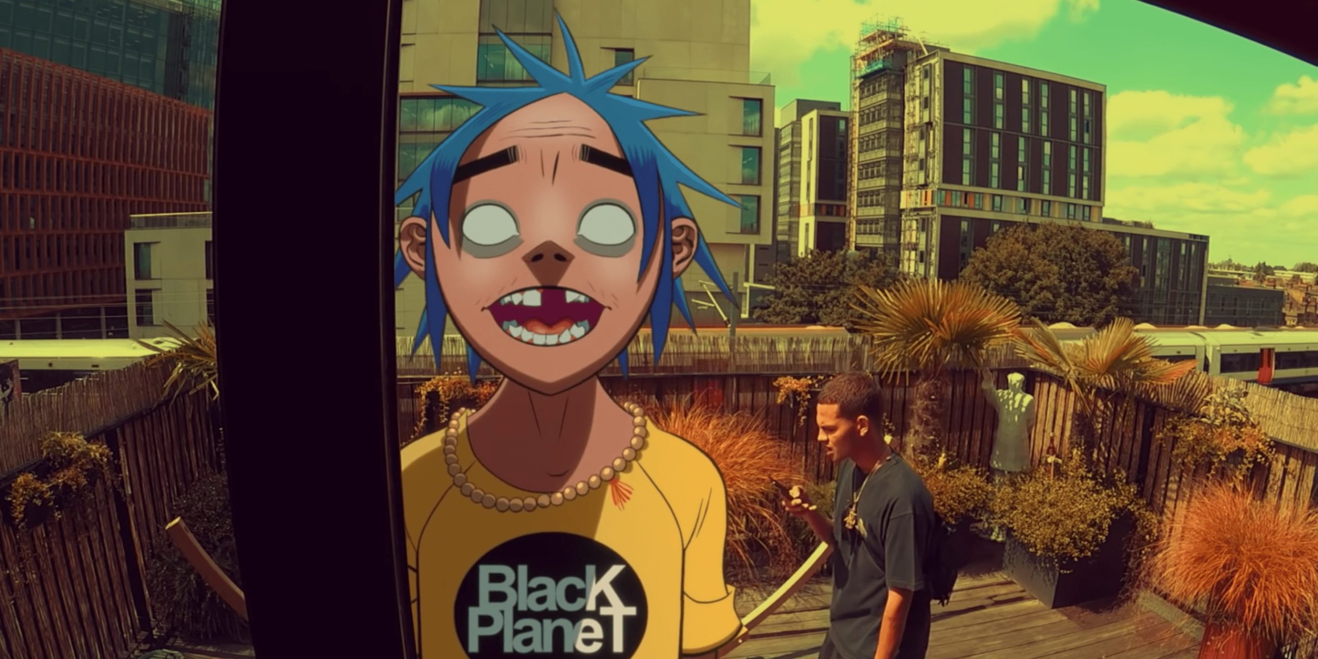 2D faces the camera in a yellow shirt that says, "Black Planet," as he sings outside on the patio in an urban city. Slowthai paces behind him. 