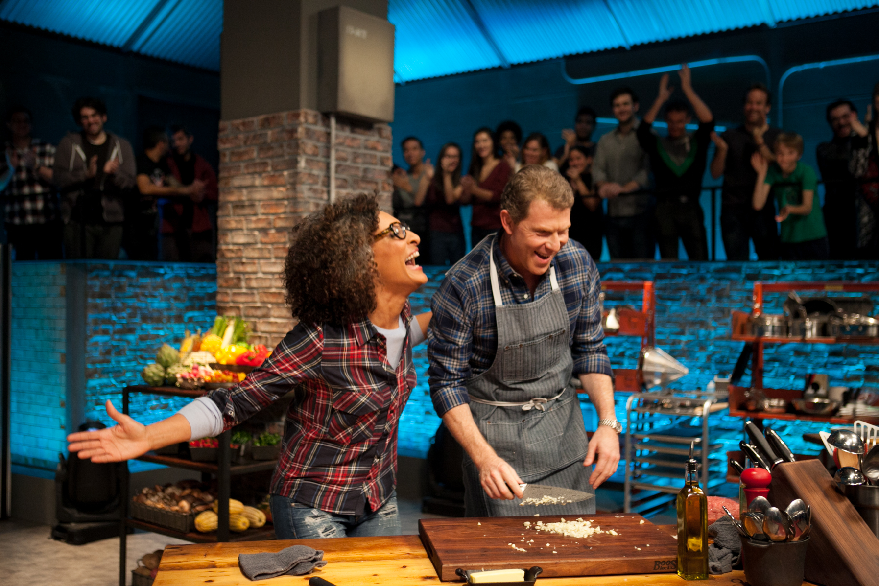 Bobby Flay stands in front of his competition station cooking, while woman stands laughing beside him. 