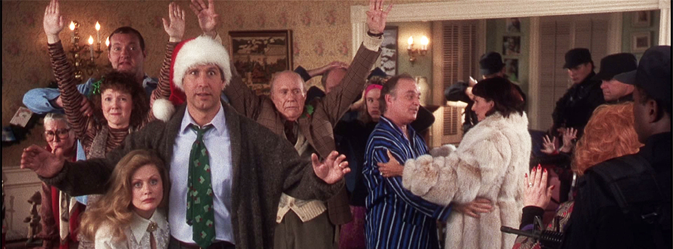 An eccentric-looking family stand in a room decorated for Christmas, all in various poses.