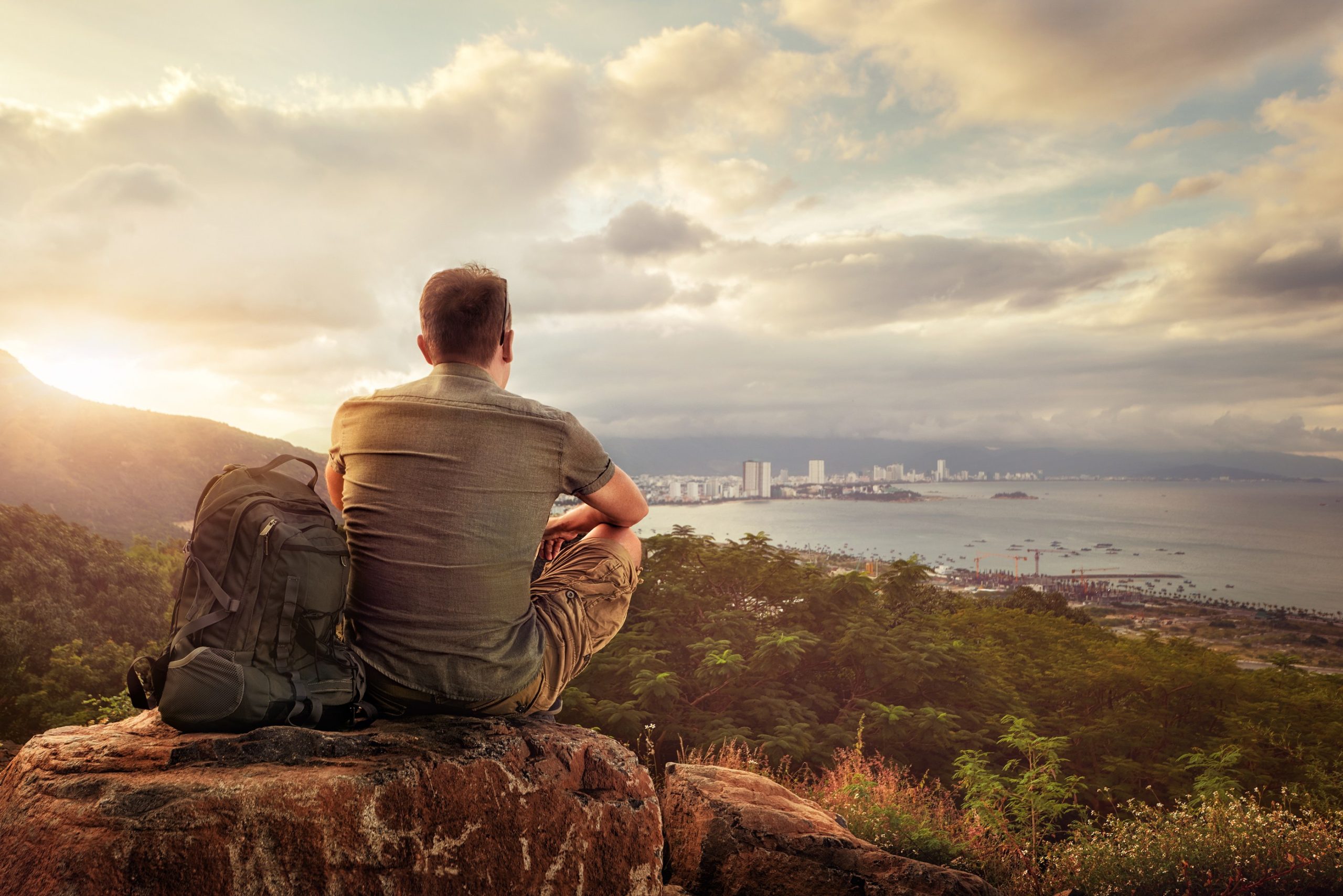 Man with backpack sits on a mountain and stares out at the water and land below.