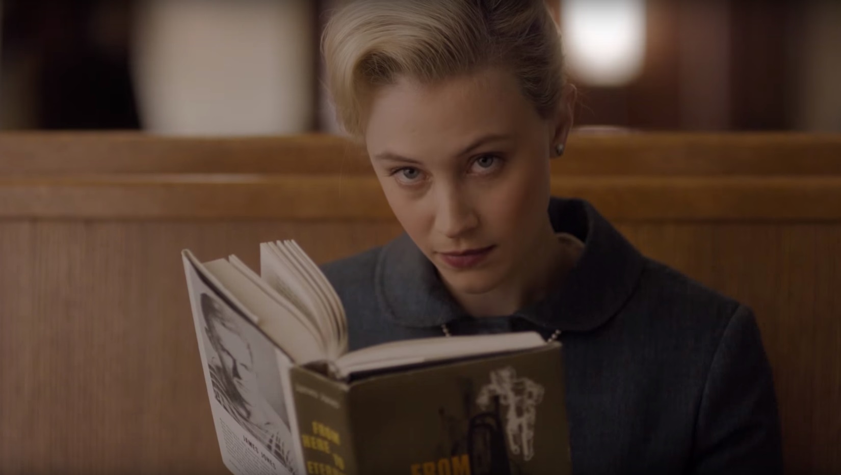Sadie reads a book and stares at someone off-camera.