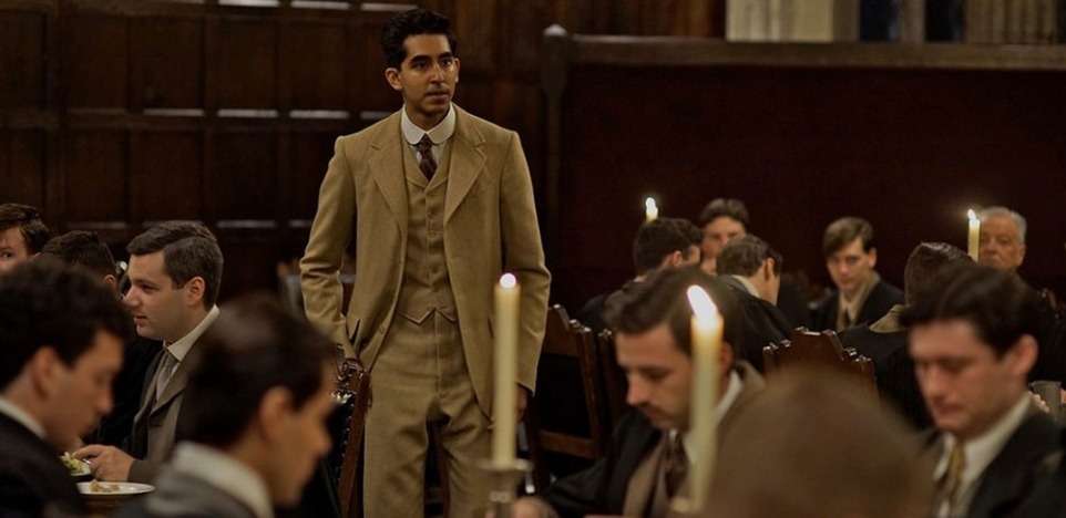 Ramanujan, dressed in a light brown suit, stands up in a room of men dressed in black suits, all sitting. 