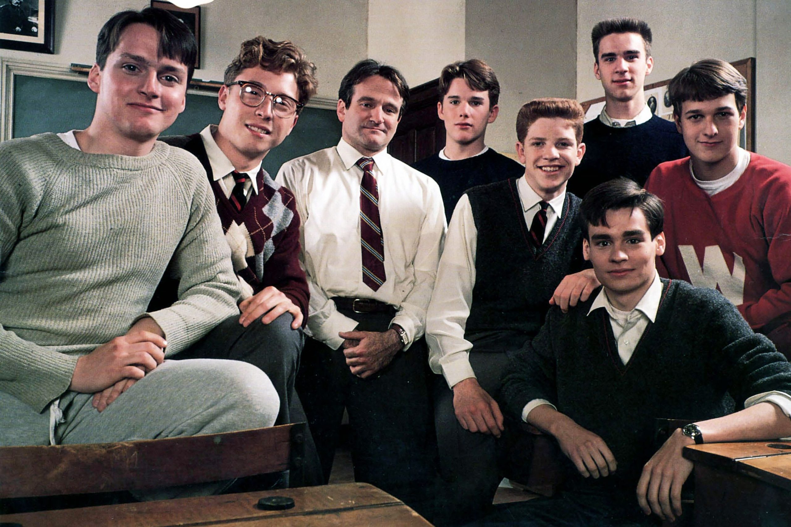 A group of male students sit perched around their teacher in a classroom, all facing the camera,