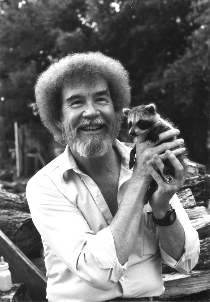 A close-up of Ross smiling in the outdoors while holding a baby raccoon.