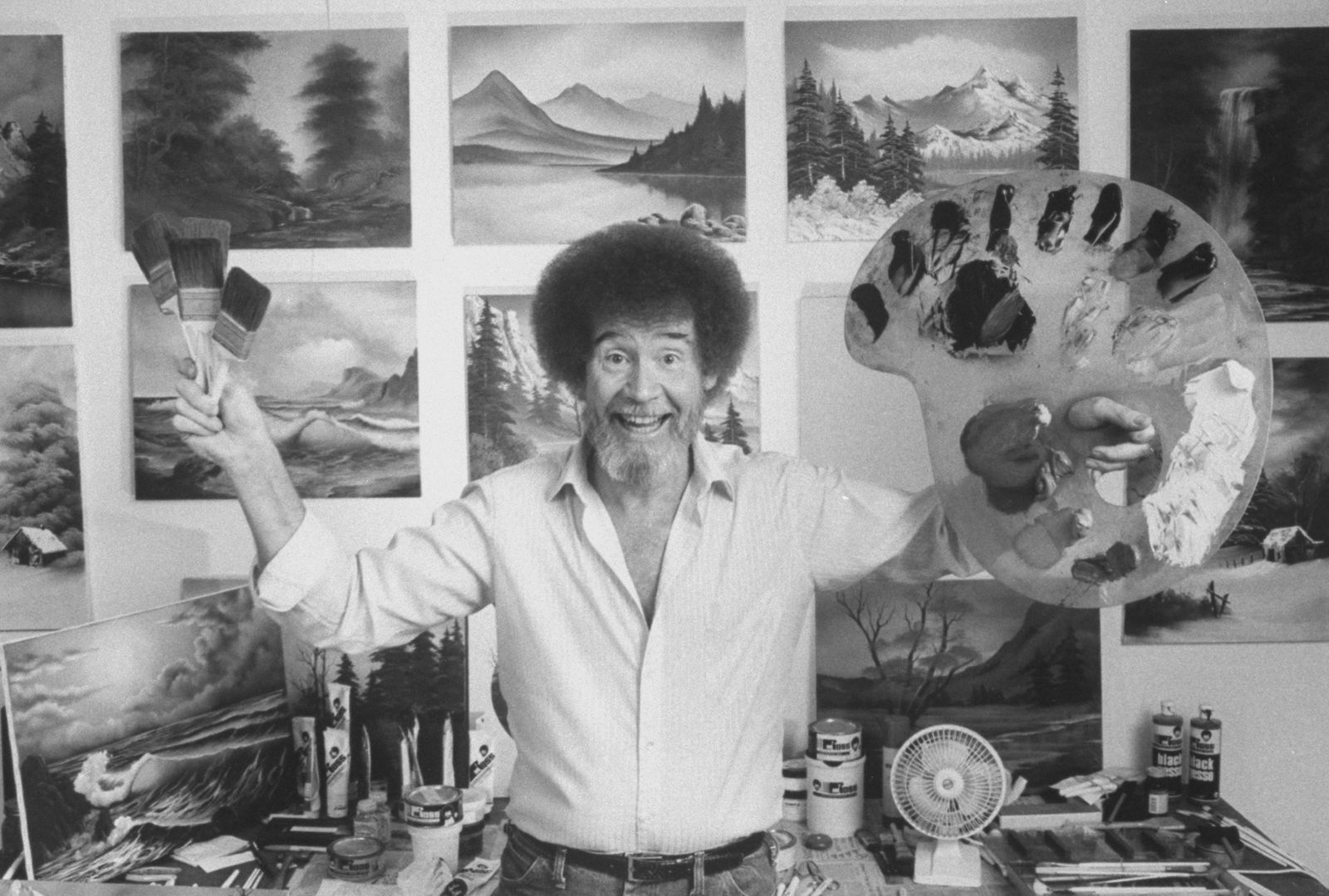 Bob Ross smiles at the camera while holding his paint brushes and pallet in front of a wall of his finished canvases.