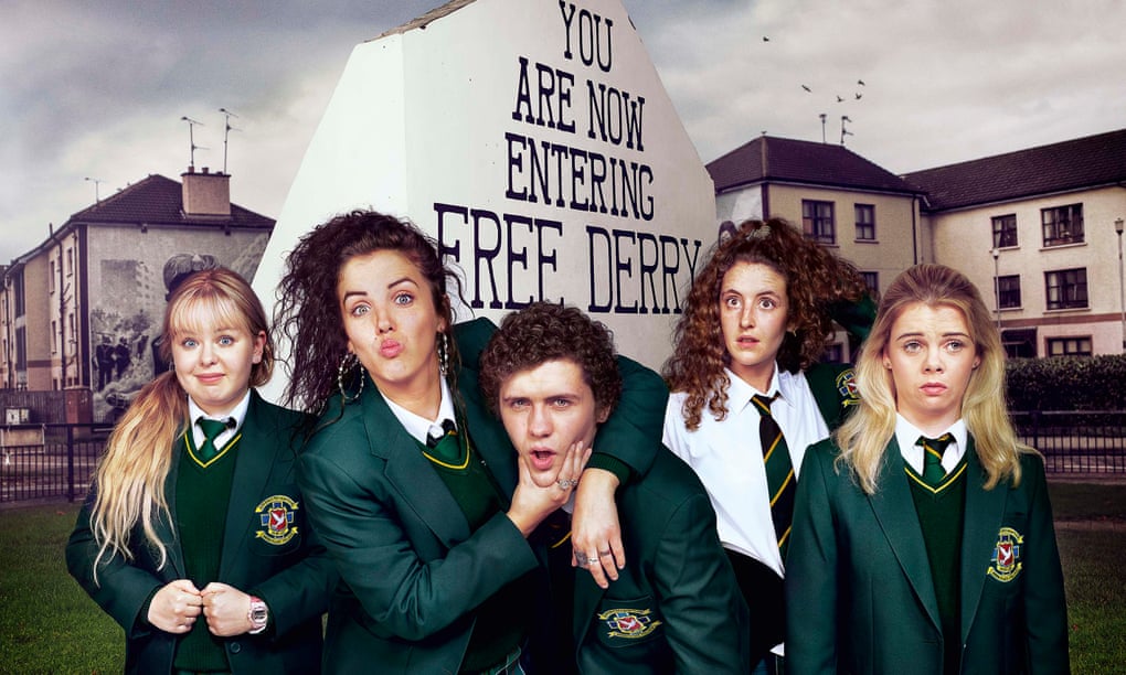 The Derry Girls stand in a row facing the camera as Michelle pinches James' cheeks.  They are in front of a sign that says 'You are now entering Free Derry'.