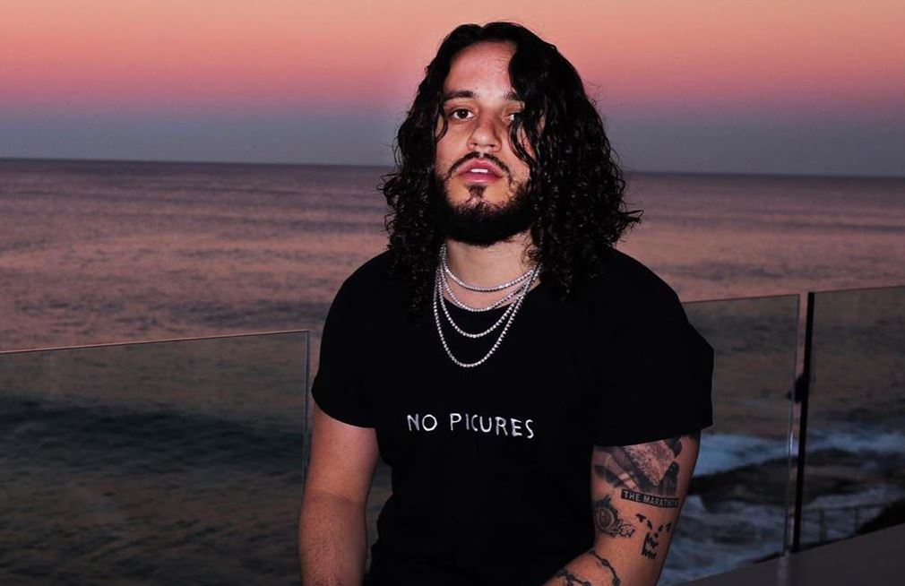 Russ stands in front of a beach and a sunset while wearing a shirt  that reads "No Pictures."