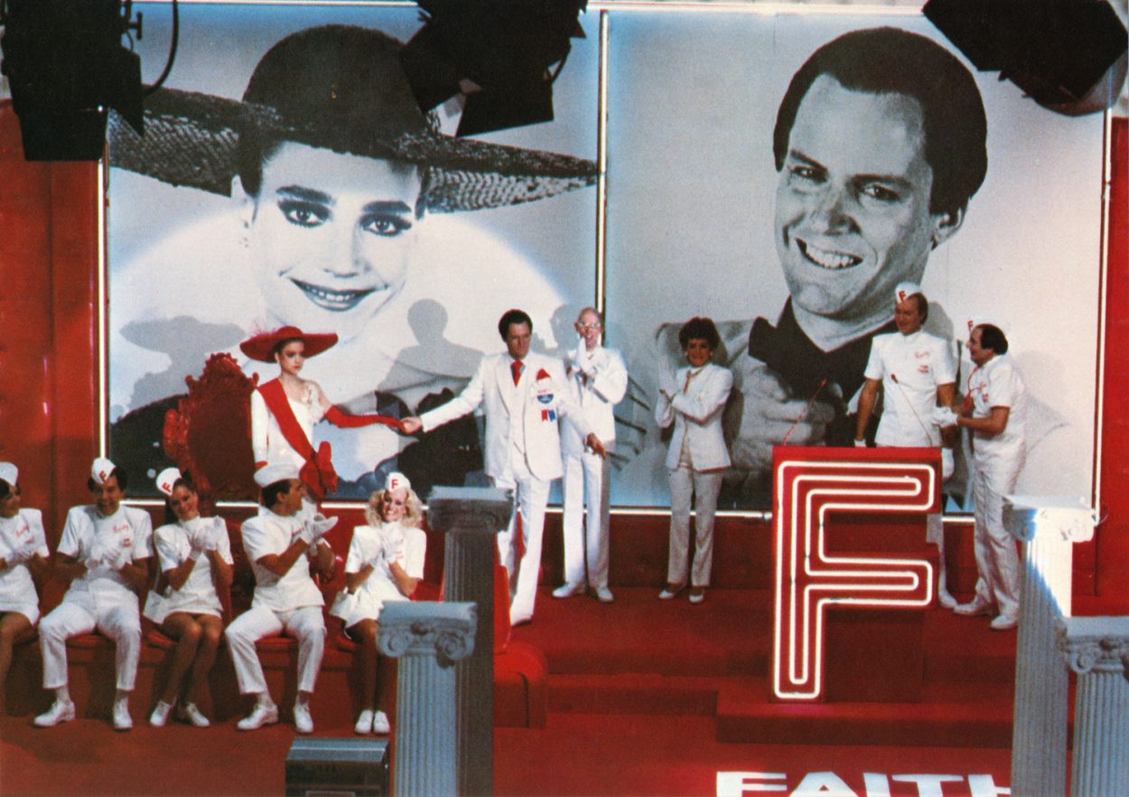 Janet and Farley are standing on a red stage with gigantic images of Janet's and Farley's faces behind them.