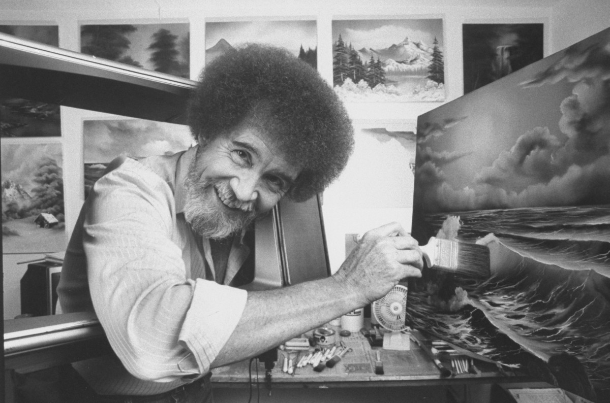 Bob Ross smiles at the camera while painting an ocean landscape; the photo is monochromatic.