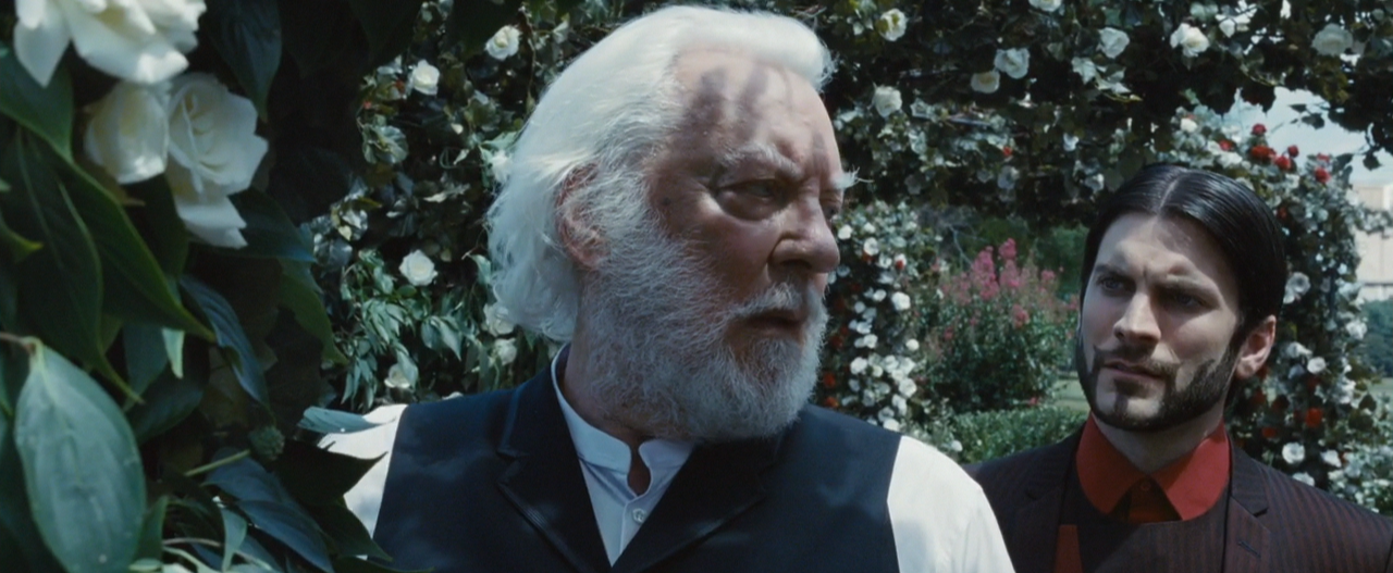 President Snow (Donald Sutherland) wears a white rose lapel in his garden of white roses while Seneca Crane (Wes Bentley) looks at Snow from behind.