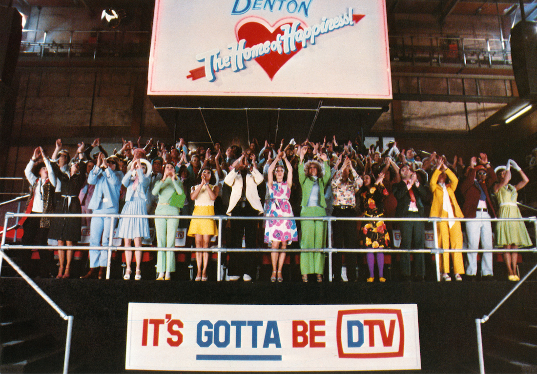 Denton TV's live studio audience in Shock Treatment is applauding. There is a sign that says, "Denton: The Home of Happiness," and a sign that says, "It's gotta be DTV."