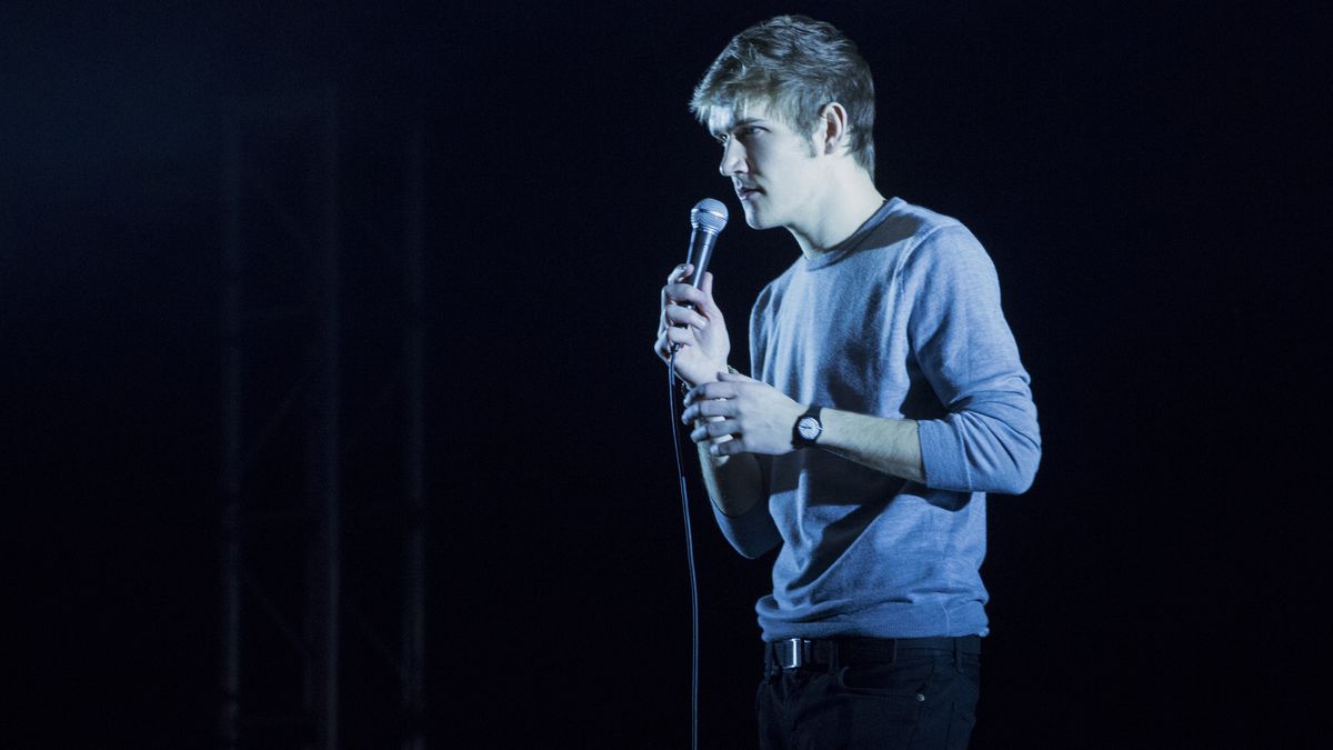 Bo Burnham stands on a dramatically lit stage with a microphone in his hand.