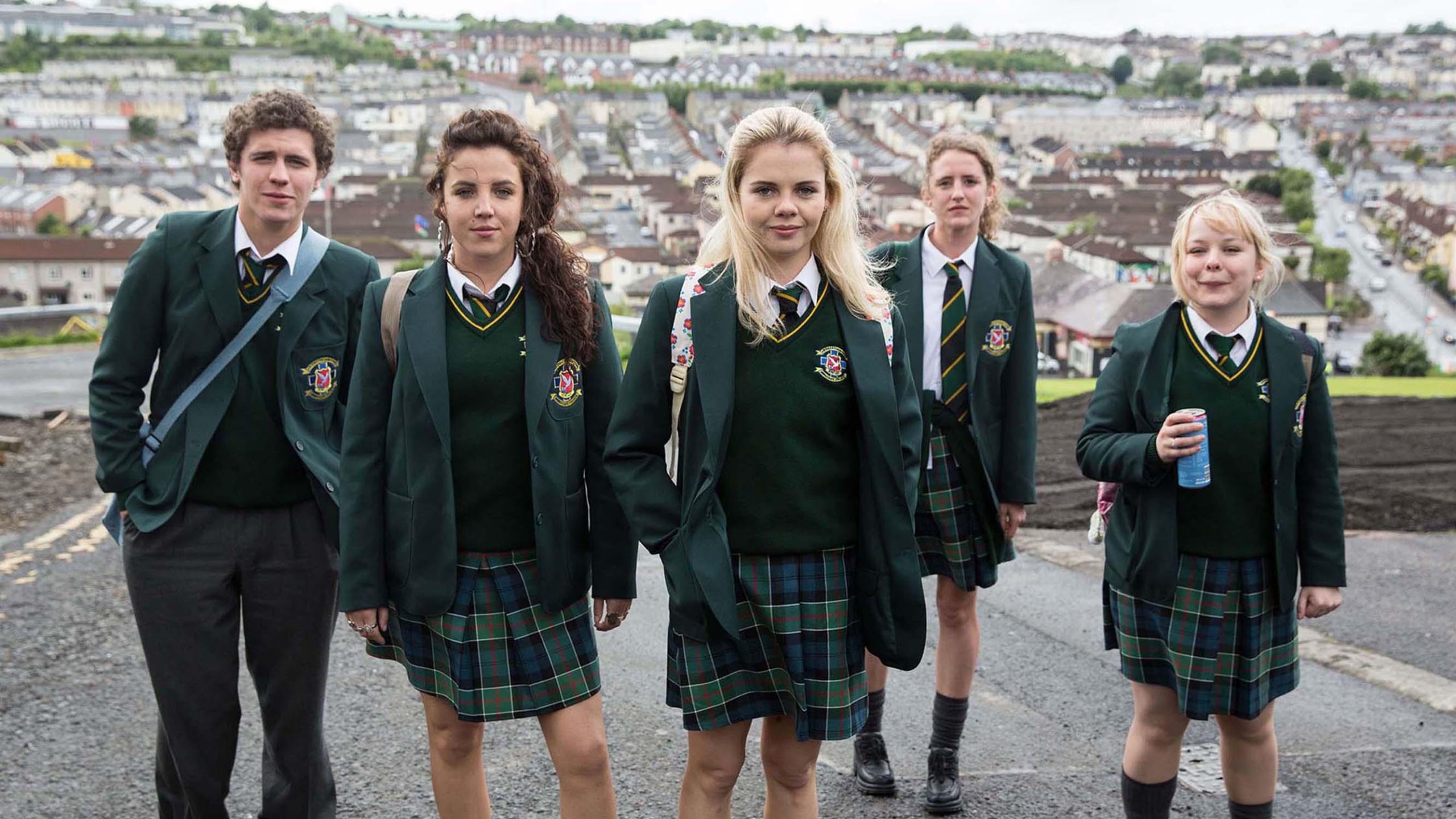 The Derry Girls stand in a line on a paved road.  Erin is standing at the forefront of the group while Clare, to her side, is holding a canned drink.