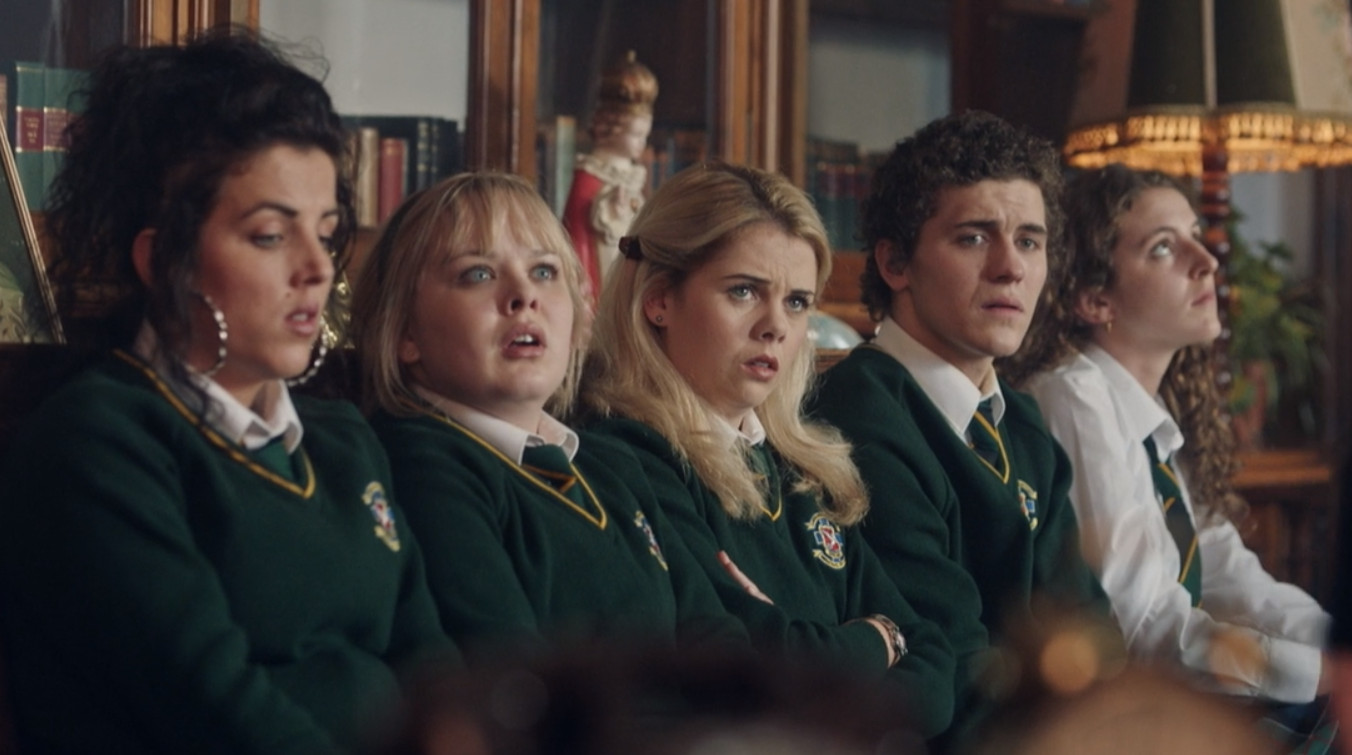 The Derry Girls sit next to each other on a bench.  They have pensive expressions on their faces, suggesting that they are in trouble.