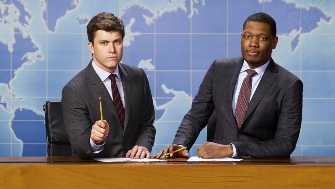 Weekend Update anchors Colin Jost and Michael Che pose at their iconic desk. 