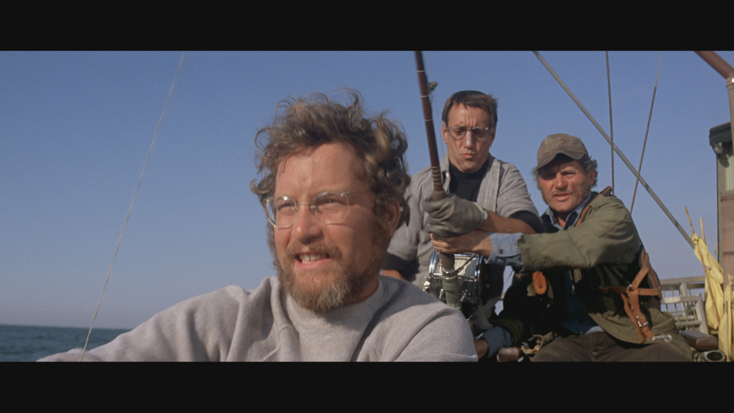 The three men stare at the shark in the distance, two of them holding a fishing line.