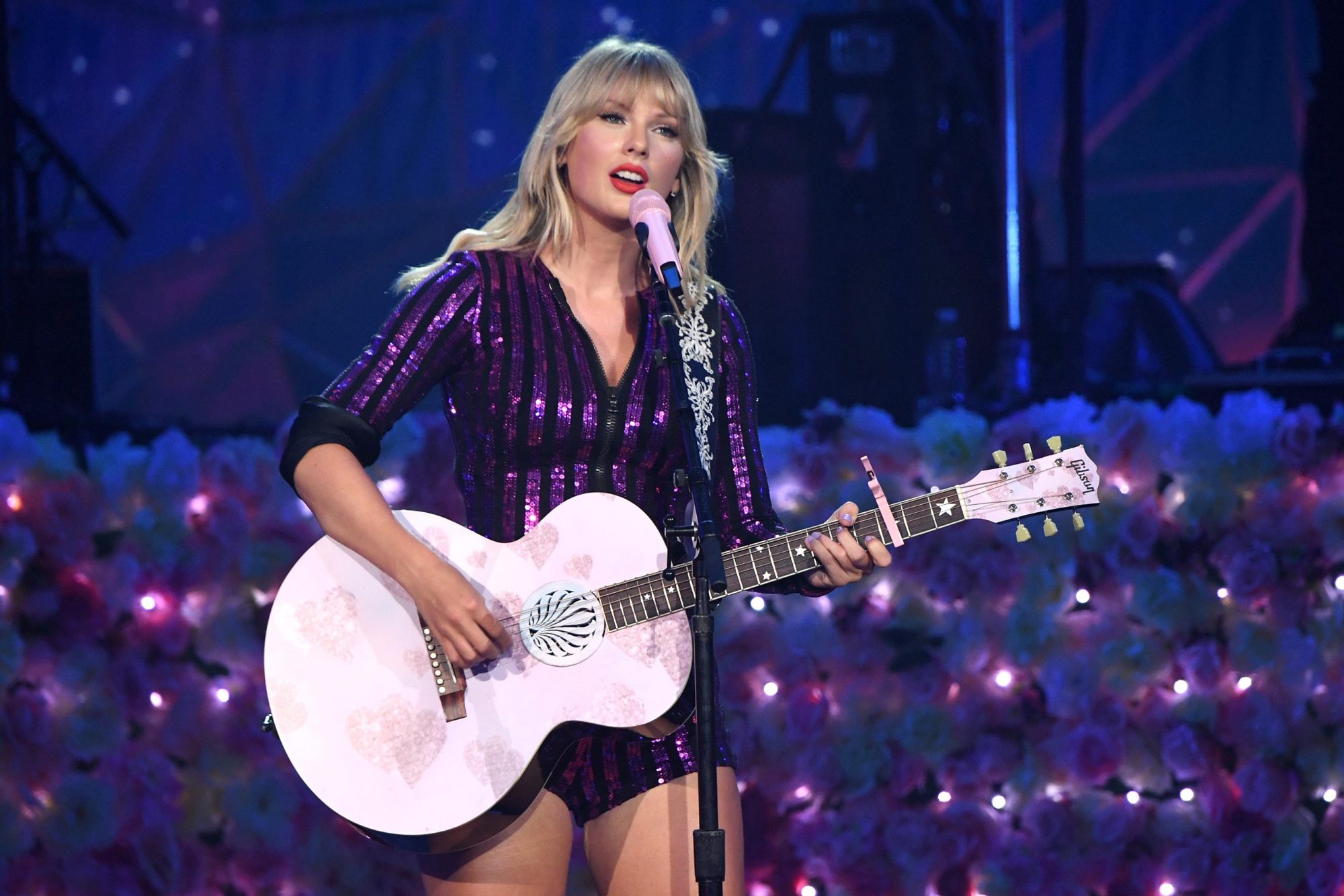 Swift stands at a microphone, holding a pink guitar and singing.