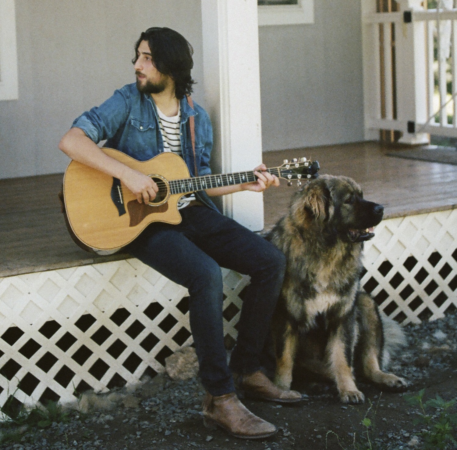 Noah Kahan sits outside on the porch with his dog while holding his guitar.