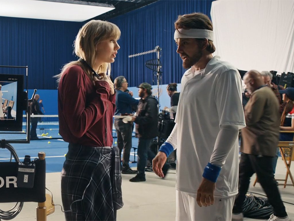 Taylor Swift directing the music video for "The Man." She stands in headphones, talking to her male counterpart, who looks like Swfit in all-white tennis attire and with facial hair.