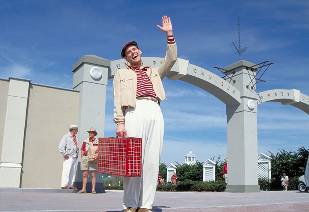 Truman Burbank from The Truman Show stands holding a suitcase and waves.