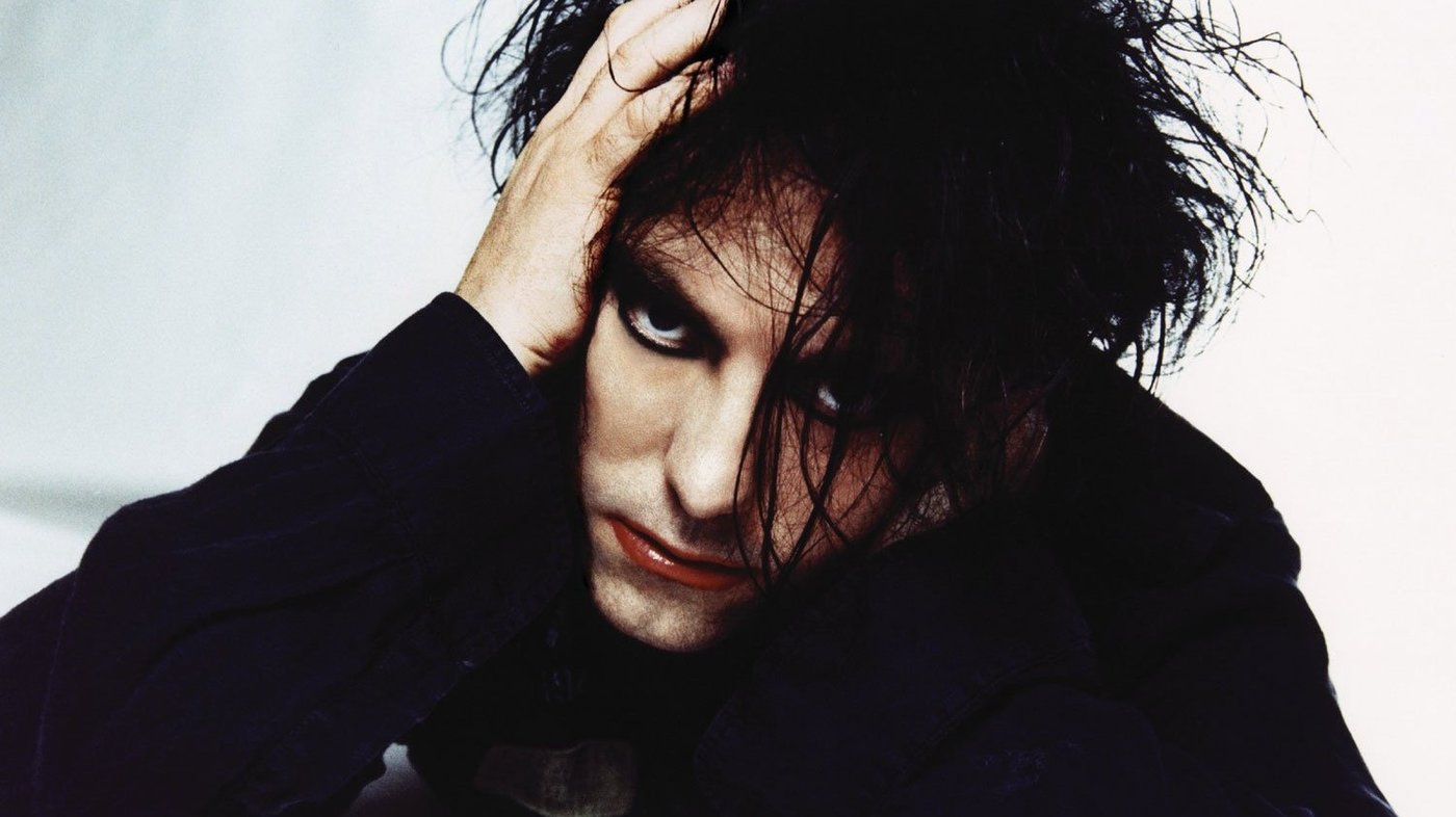 The lead singer of The Cure, Robert Smith, looks at the camera with his hair in his eyes and one hand on his head.