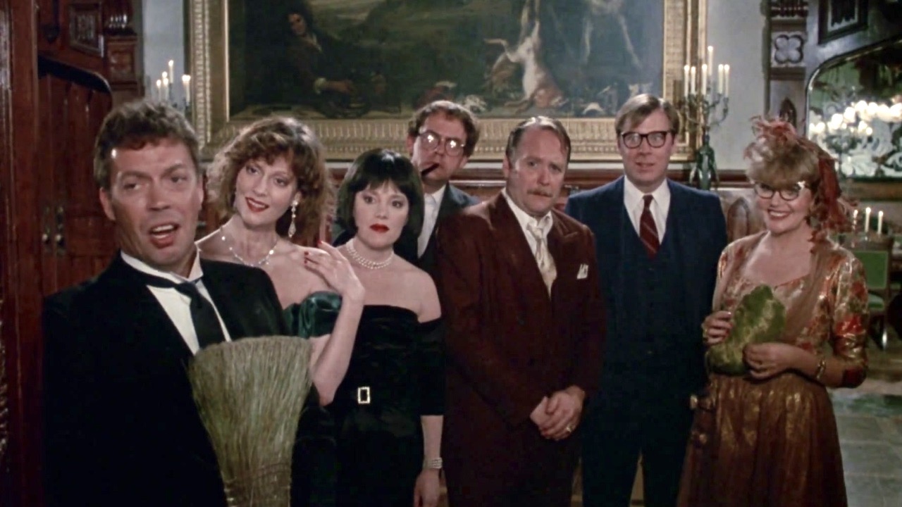 The cast of Clue stands looking out the door of the mansion. Wadsworth holds a broom and talks to a person outside the frame.