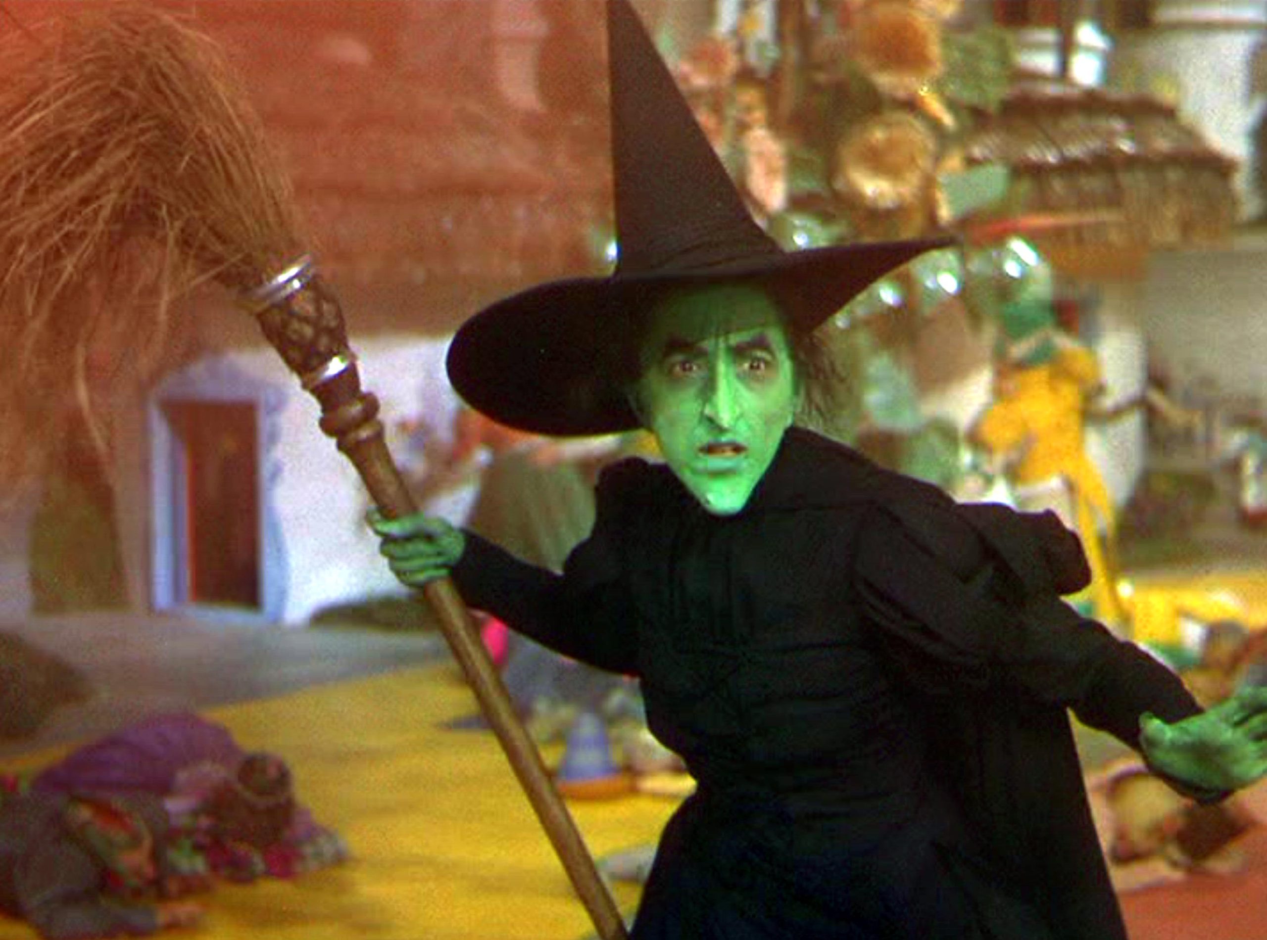 The Wicked Witch of the West from The Wizard of Oz stands on the yellow brick road, holding her broomstick.