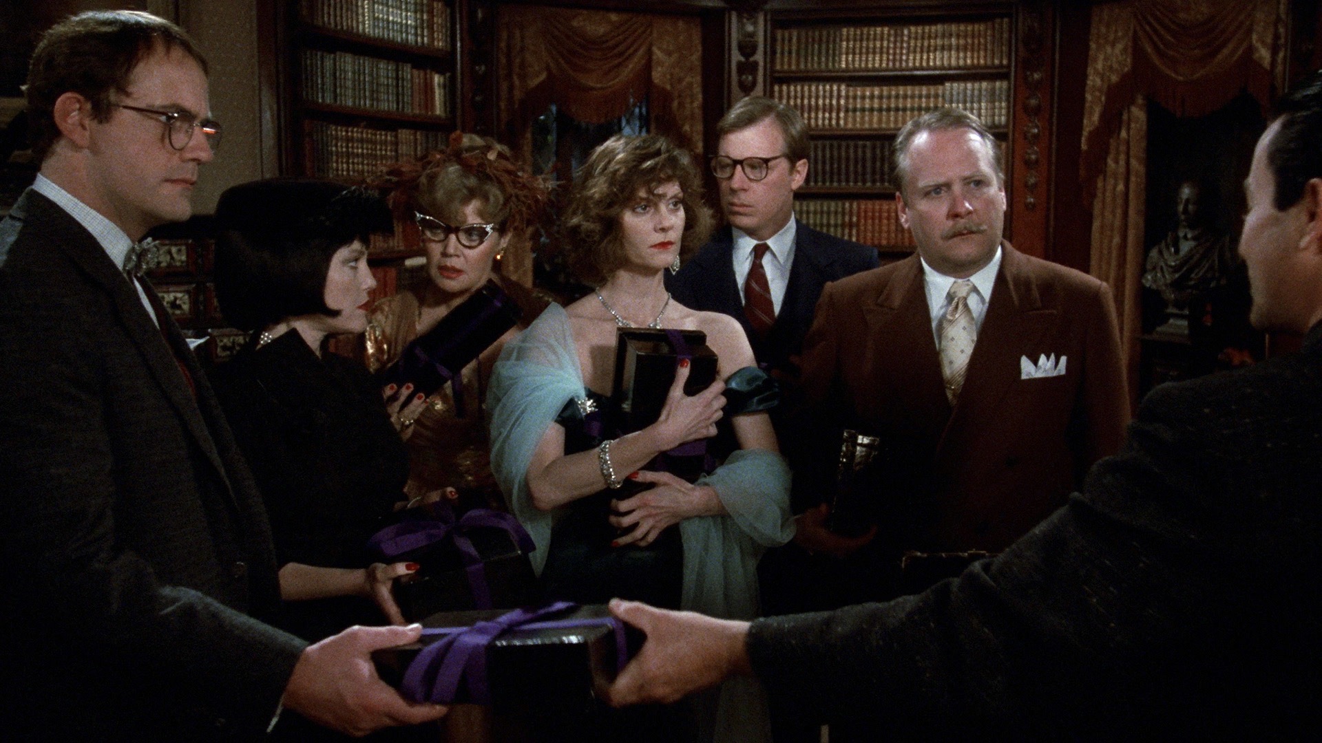 Mr. Boddy hands Professor Plum a black package tied with a purple ribbon. Colonel Mustard, Miss Scarlet, Mr. Green, Mrs. Peacock, and Mrs. White stand in the background, all holding similar packages.