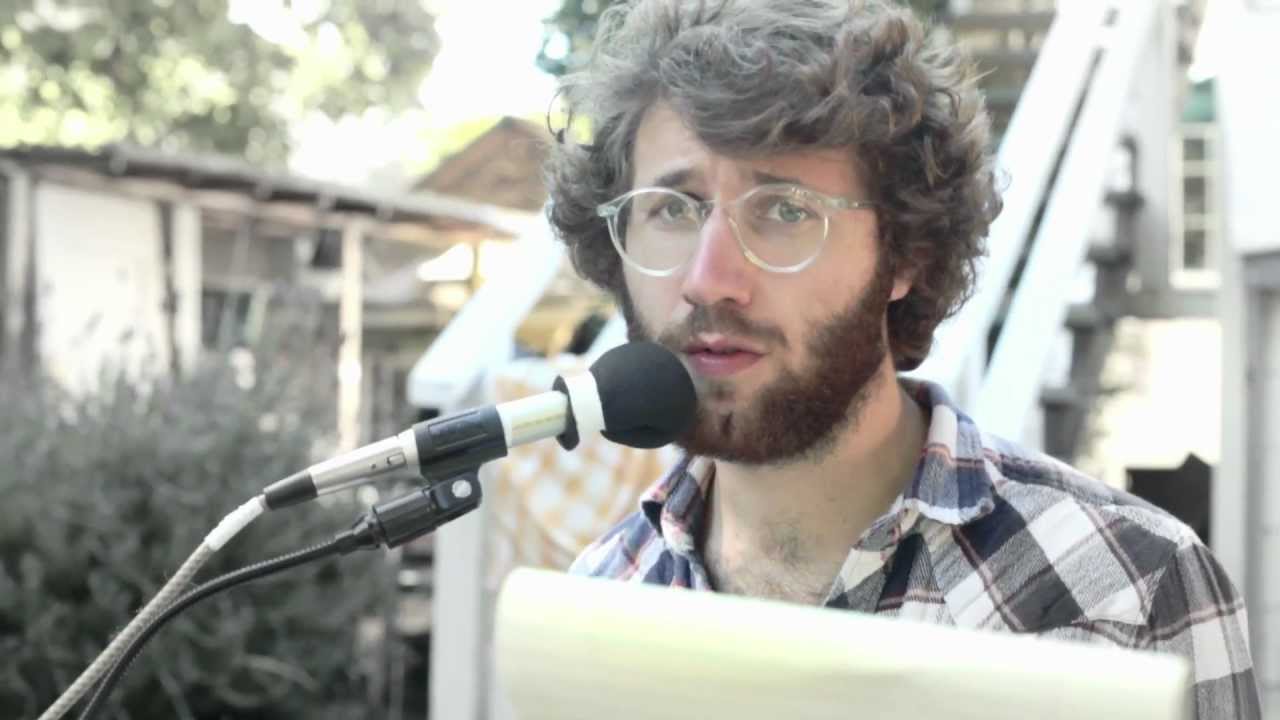 Jack Stratton as Mushy Krongold, speaking into a microphone and lookin wistfully at the camera. 