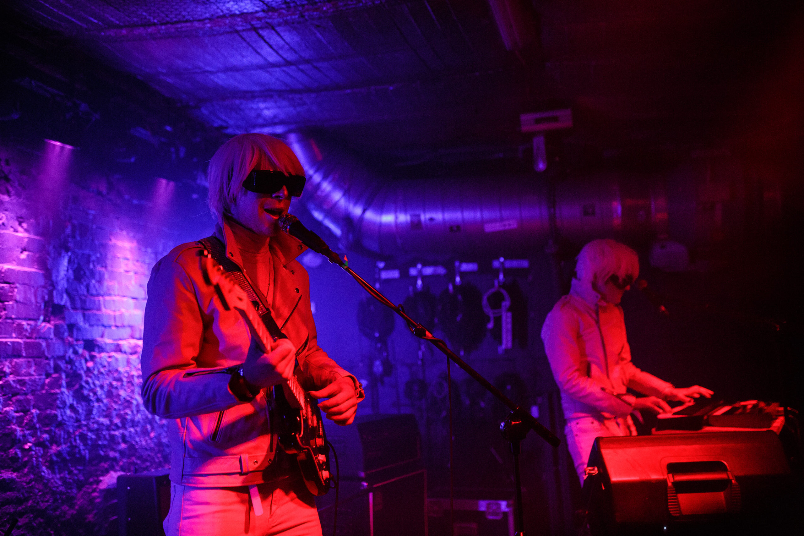 Deb Demure (Andrew Clinco) and Mona D (Alex Nicolaou) of Drab Majesty stand playing their music live. 