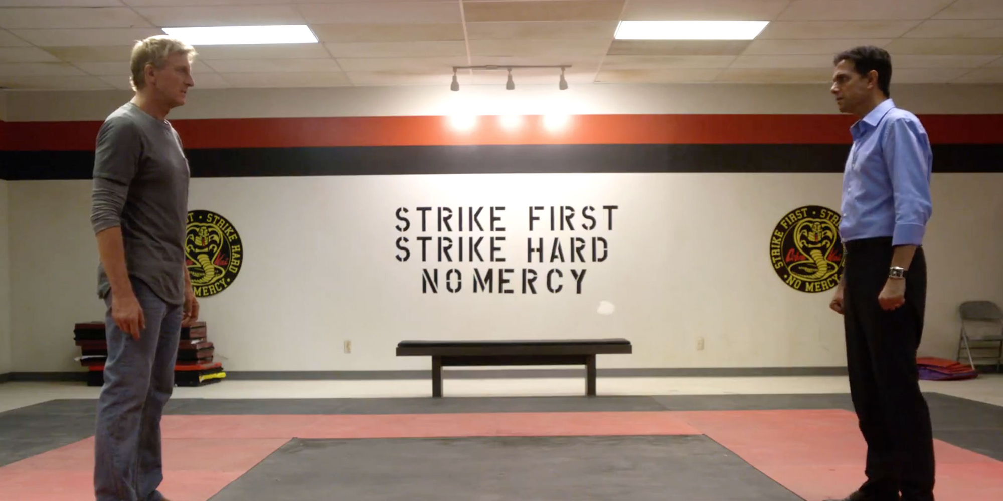 Johnny and Daniel face off in the Cobra Kai dojo. Visible between them on the wall is the Cobra Kai motto: Strike first, strike hard, no mercy.