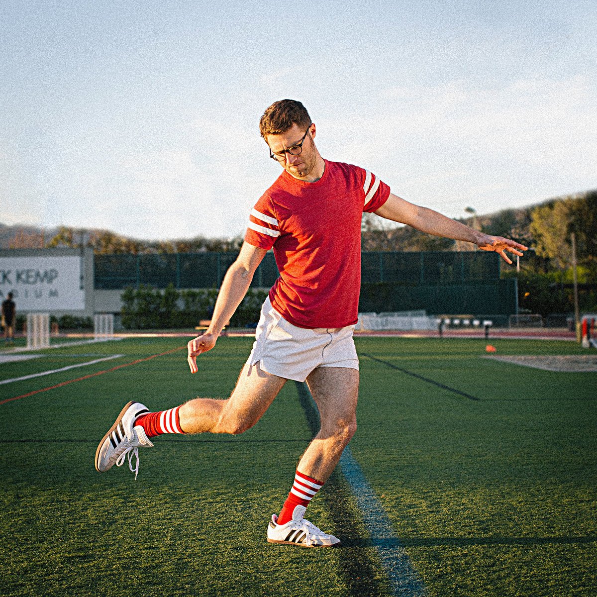 The album art for The Beautiful Game by Vulfpeck: Jack Stratton in white short shorts and a red T-Shirt, miming a kick on a soccer field. 