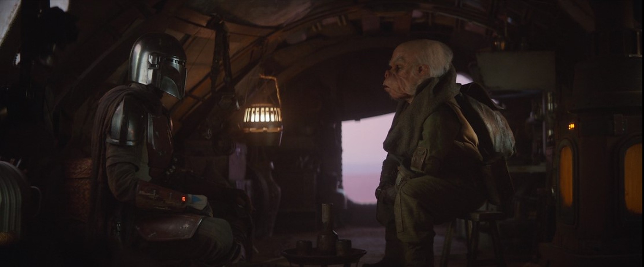 The Star Wars characters the Mandalorian and Kuiil talk in a hut.