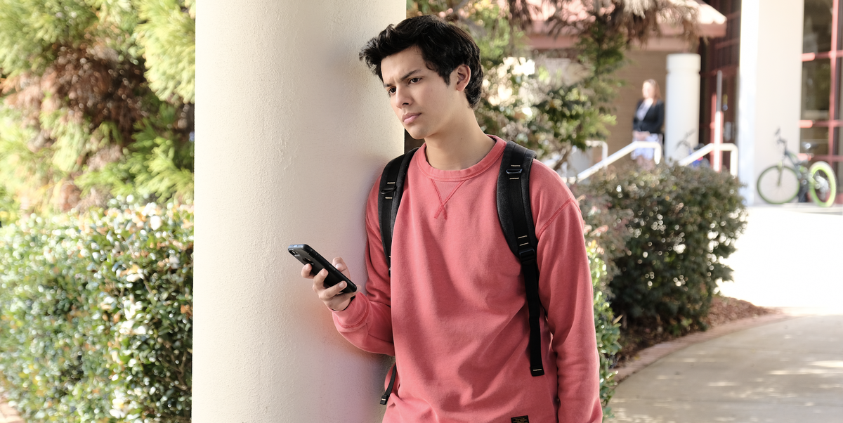 Miguel leans against a pillar holding his phone, gazing thoughtfully out into the middle distance.