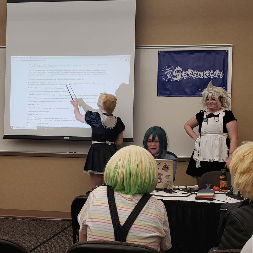 A group in cosplay and maid outfits read out fanfiction from a projector to a crowd at an anime convention.