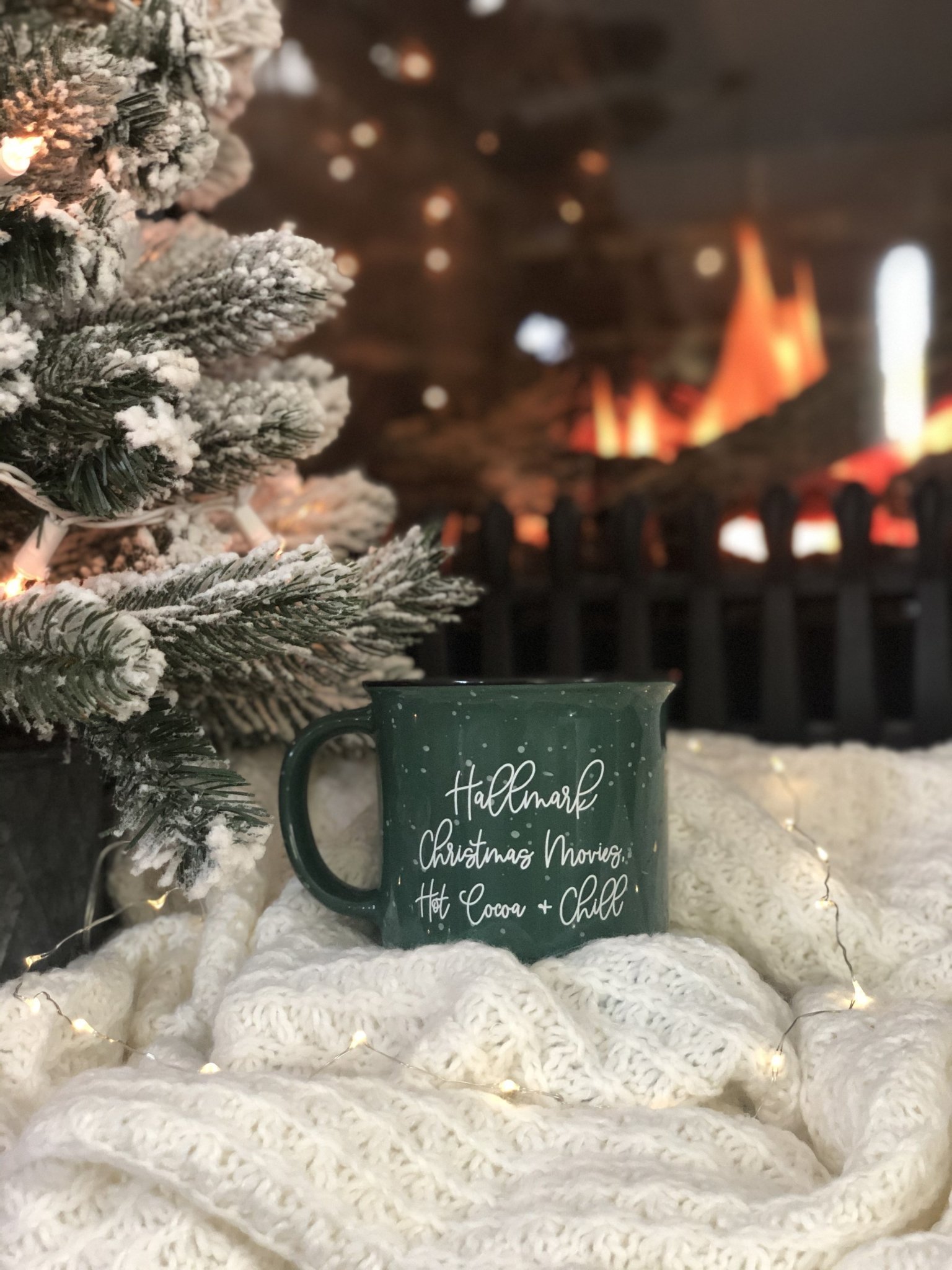 A mug that says "Hallmark Christmas Movies, Hot Cocoa & Chill" sits on a blanket in front of a Christmas tree and fireplace.