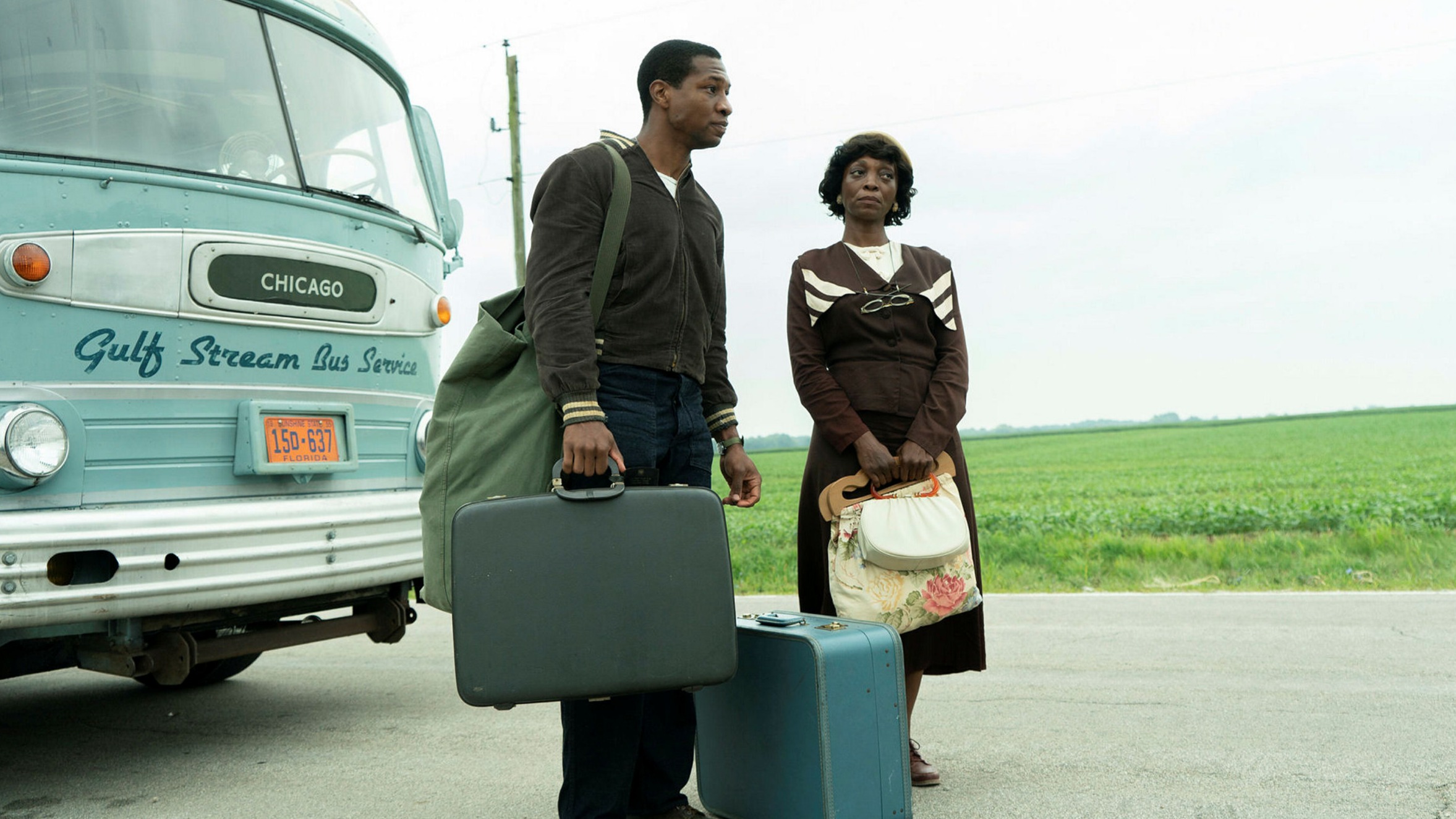 Atticus and a fellow passenger stand in front of the bus with their luggage. 