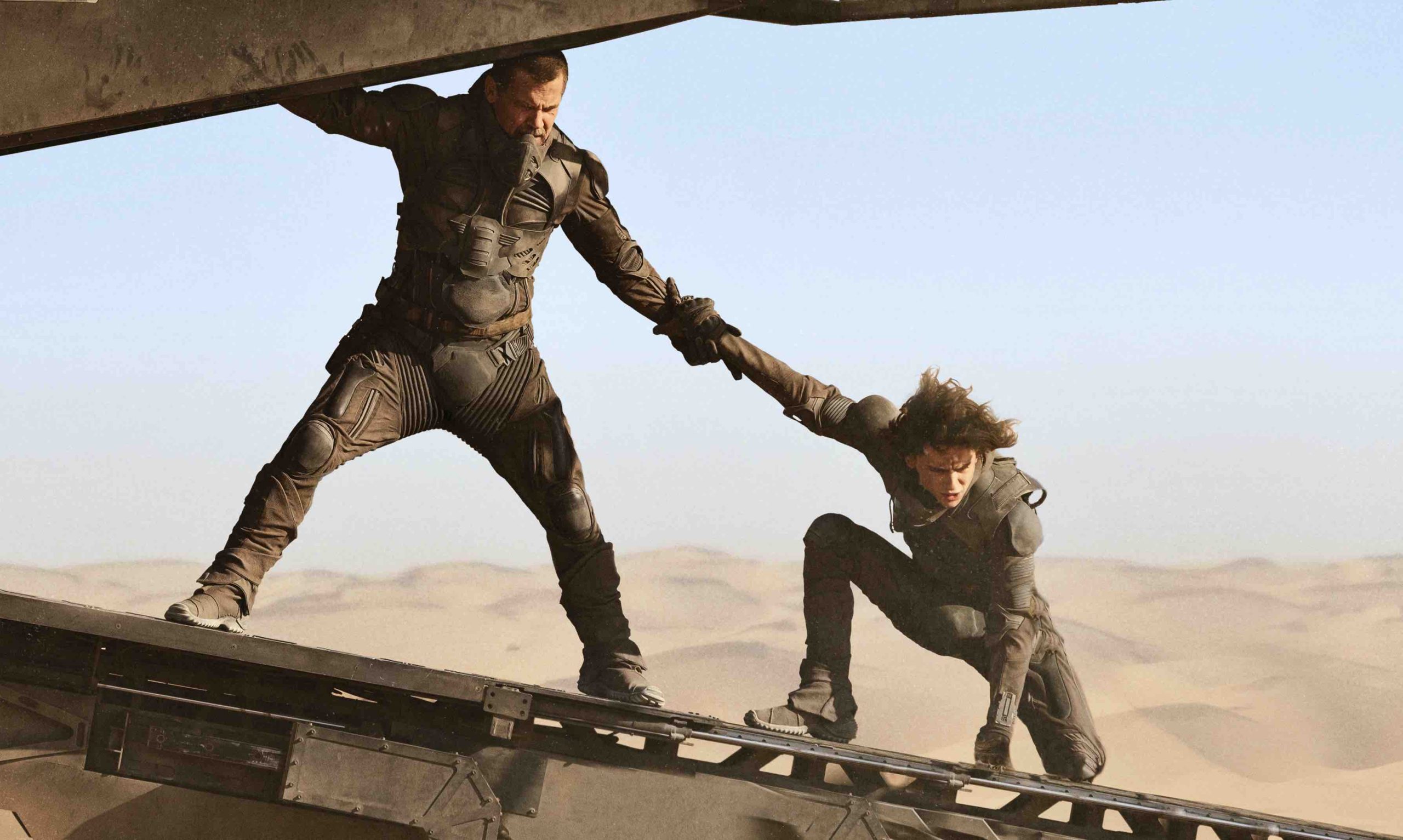 Two men stand on the ramp to an airship. In the background are endless sand dunes.