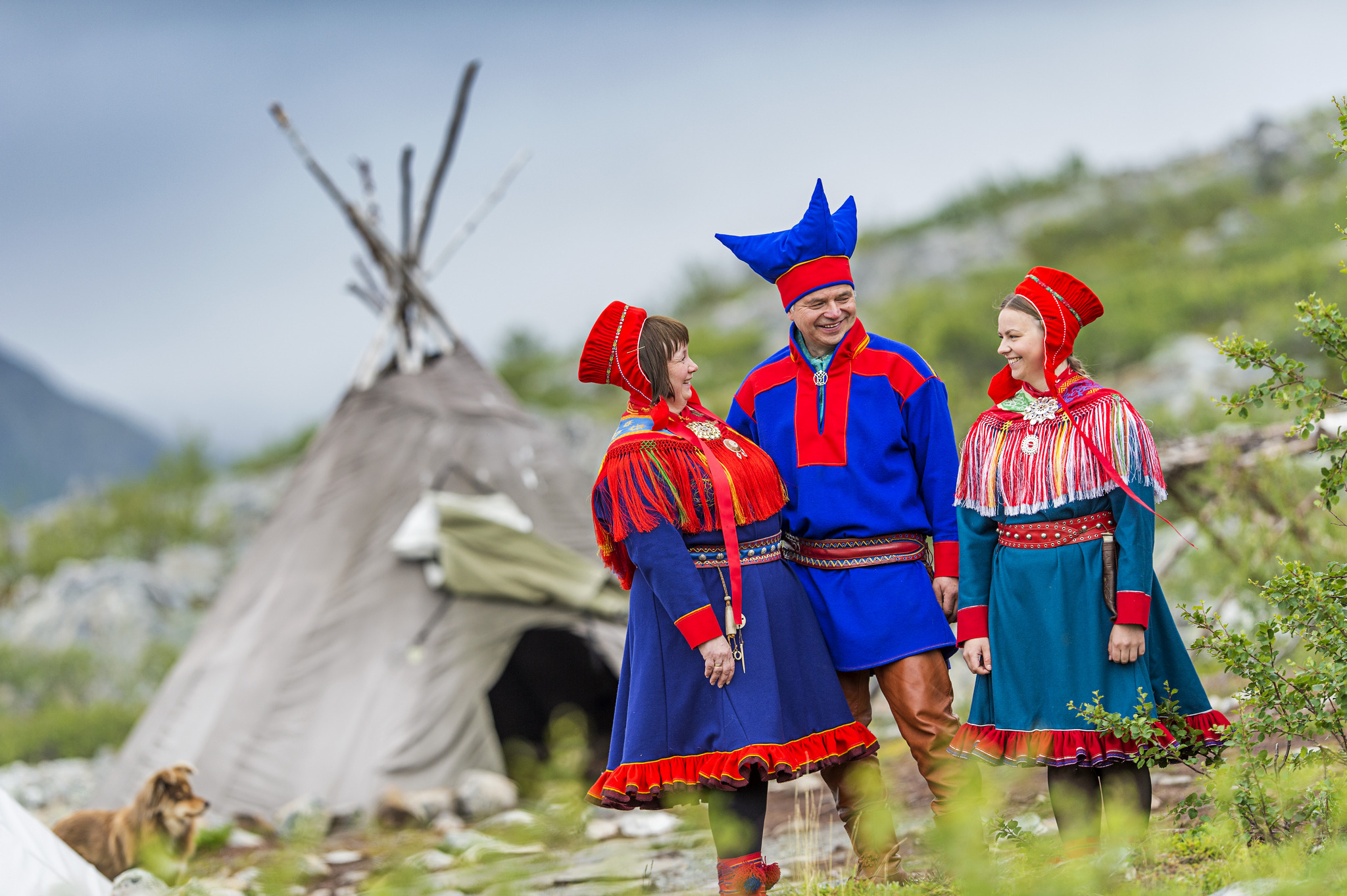 Sámi people in traditional dress stand outside what is probably a lavvu.
