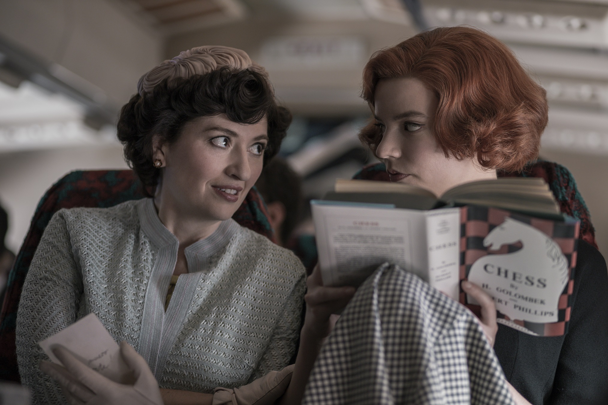 Mrs. Alma Wheatley (Marielle Heller) and Beth Harmon (Anya Taylor-Joy) sit next to each other in an airplane and engage in a conversation, while Beth attempts to read a book about chess.