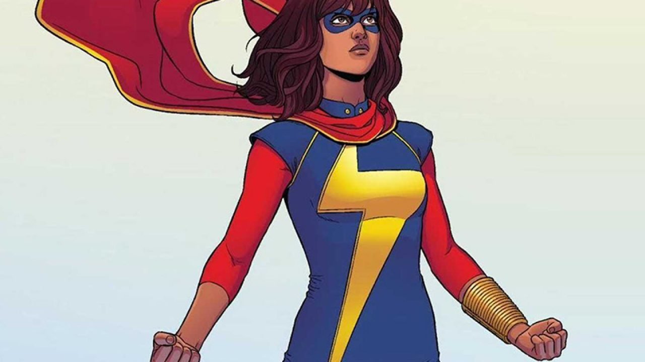 Ms. Marvel in her superhero costume and posing heroically. Her outfit is blue and red with an extended lighting bolt down the blue of her torso. She dons a blue domino mask. 