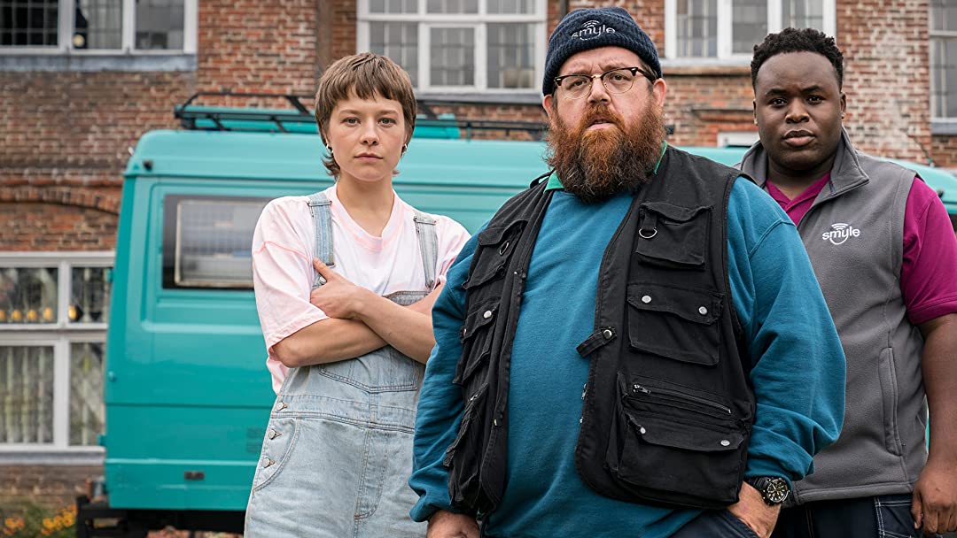 Gus stands in front of Astrid and Elton who are on either side of him. The blue Smyle broadband cable van is parked in the background.